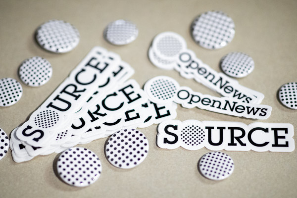 A pile of Source stickers and OpenNews dot logo buttons on a table.