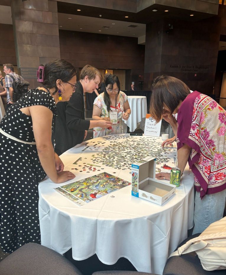 A group of five Source-con 2026 attendees gather in Memorial Hall of the McNamara Alumni Center, the conference venue, to complete a 1000 piece puzzle together at the puzzle table.